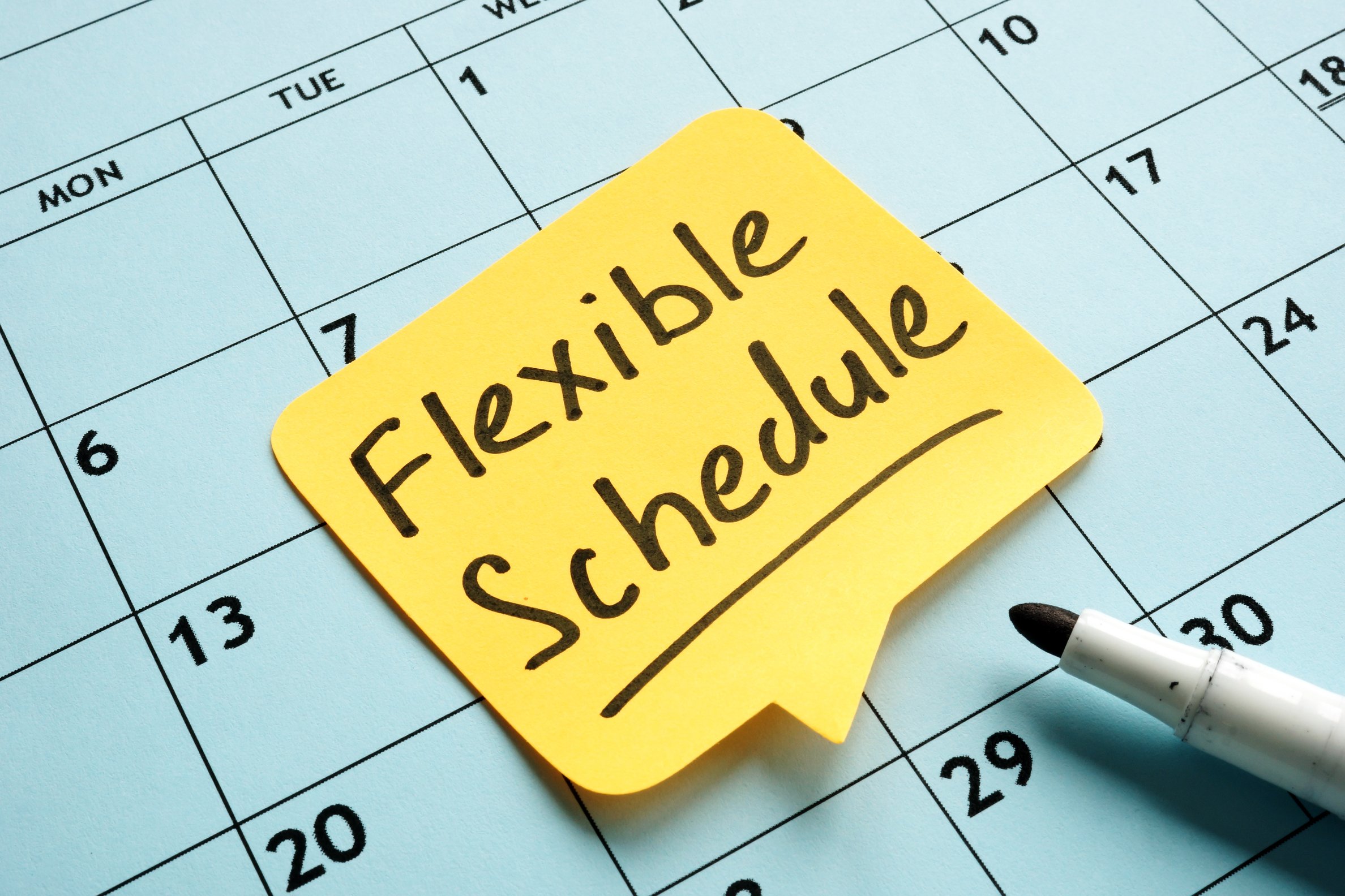  A yellow sticky note with the words 'Flexible Schedule' written in black handwriting, placed on a monthly calendar.